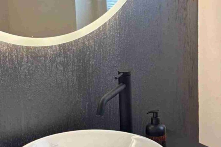 Wandfarbe FiLi di Seta Schwarzer Waschtisch mit runder Spiegelfläche und Seifenspender daneben. Dahinter eine graue Wand erstellt mit Farbe von FiLi di Seta