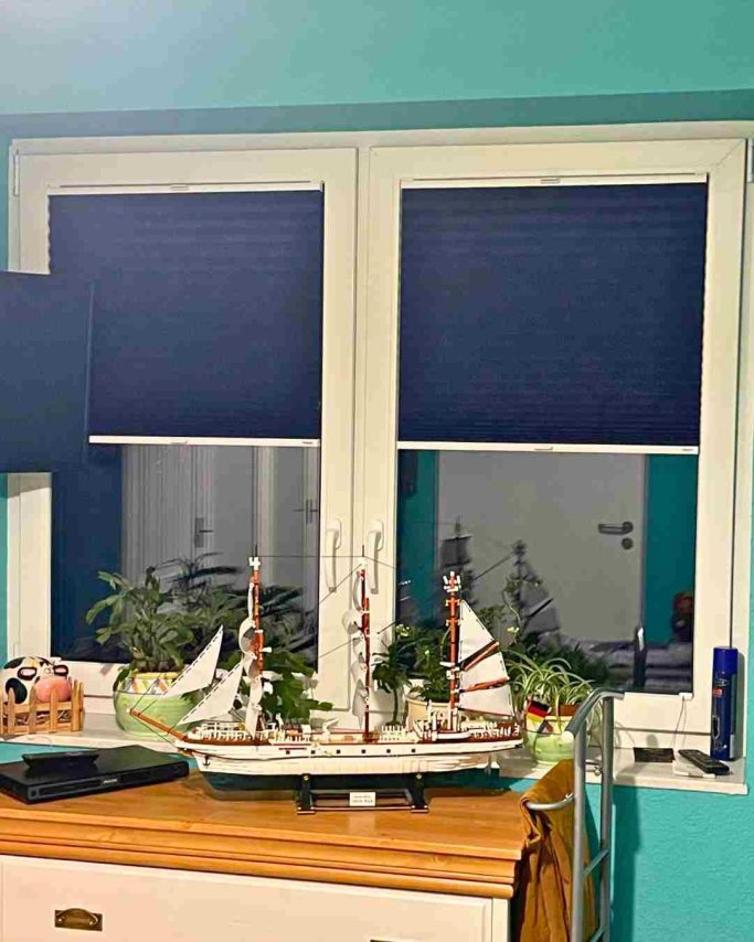 Fenster mit blauen Jalousien, einem Schiffsmodell und Pflanzen auf der Fensterbank.