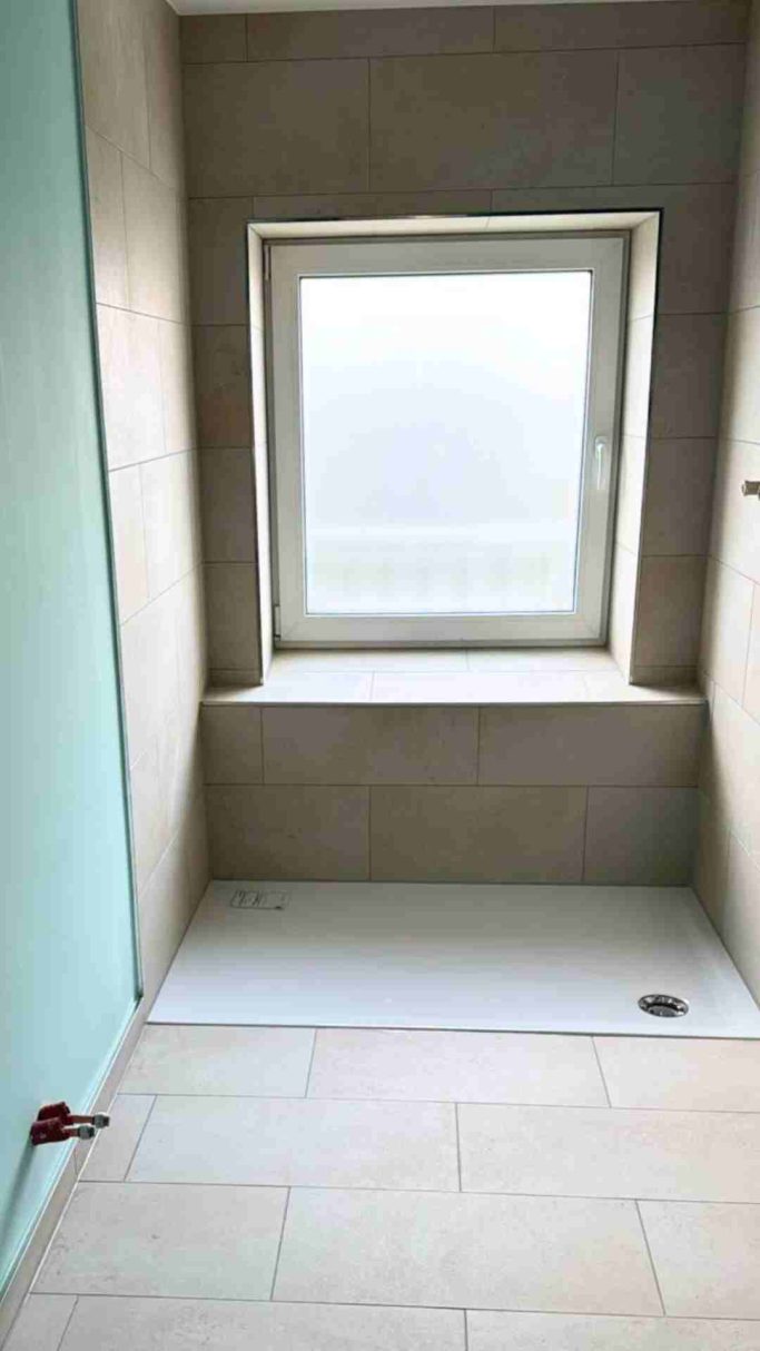 Badezimmer mit Fenster, gefliester Duschebereich und hellen Wänden.