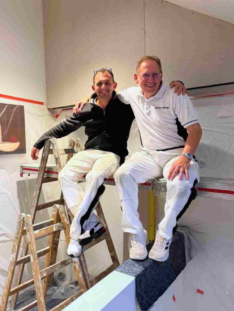 2 Mitarbeiter der Firma Raumgestaltung Michael Breuer sitzen lachend auf einer Treppe und halten sich kumpelhaft im Arm
