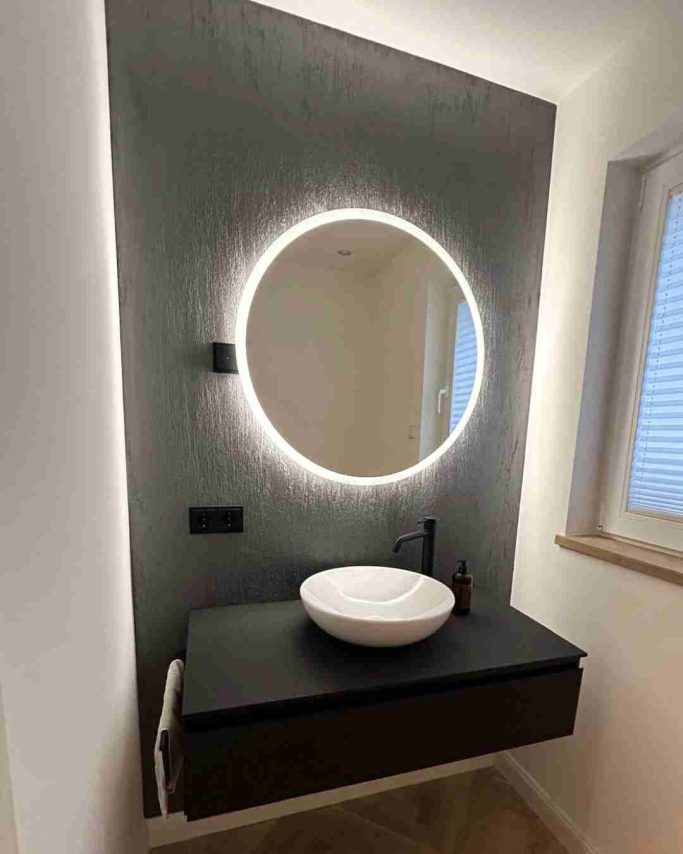 Modernes Badezimmer mit runder, beleuchtetem Spiegel und Waschbecken auf schwarzer Oberfläche.