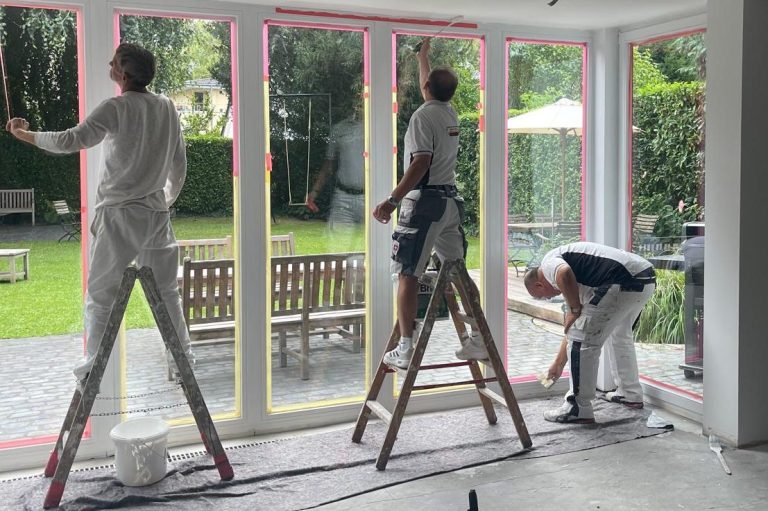 Lackierarbeiten Köln Drei Maler streichen Fensterrahmen in einem Garten mit Tisch und Stühlen.