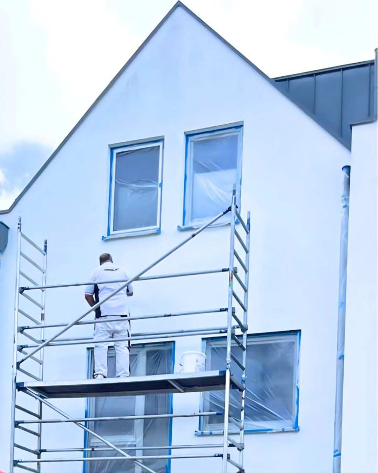 Ein Maler steht auf einem Gerüst und streicht das obere Stockwerk eines weißen Hauses in Köln.