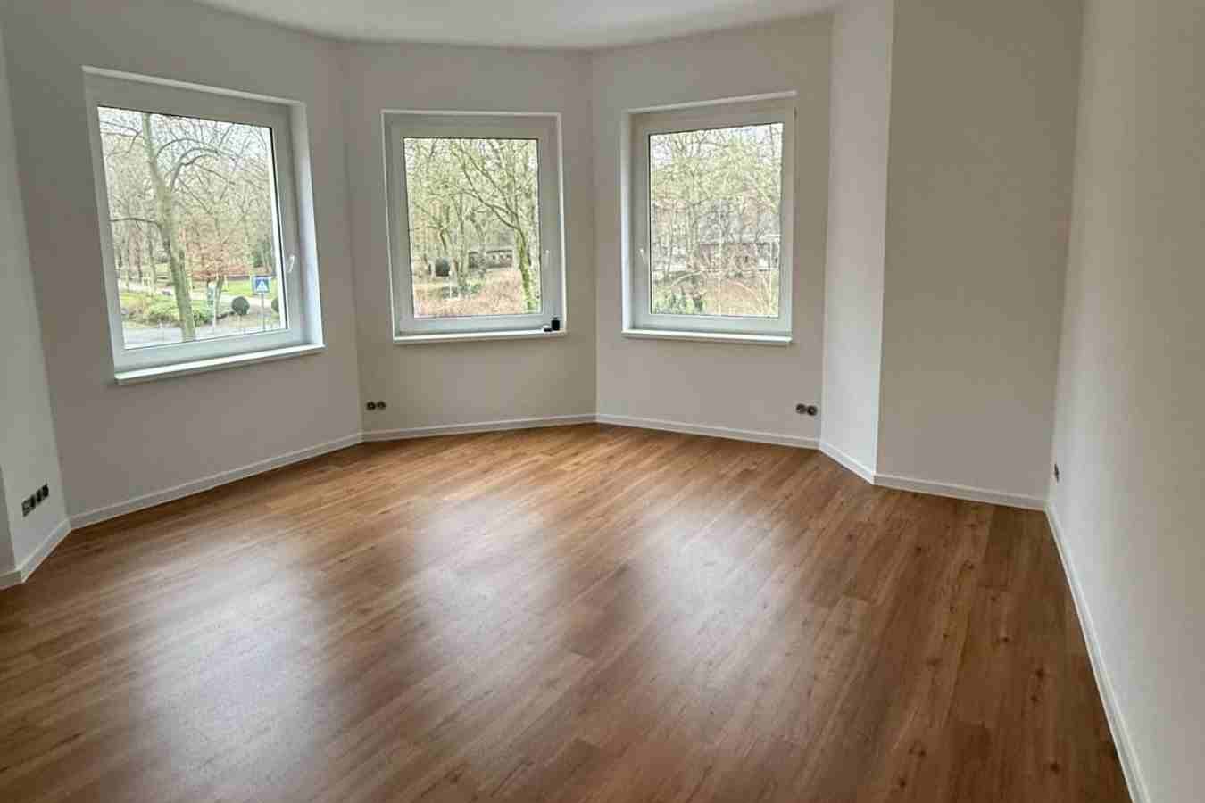 Heller, leerer Wohnraum mit Fenstern, frisch gestrichenen Wänden und neu verlegtem Holzfußboden.