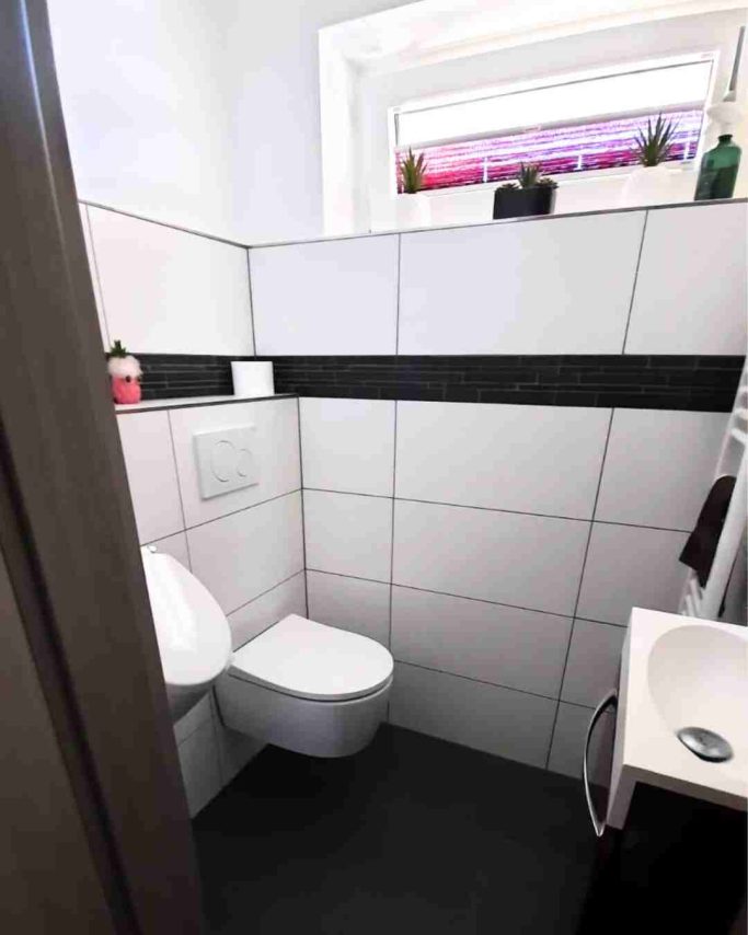 Kleines Badezimmer mit WC, Waschbecken und Fenster, halber gefliest.
